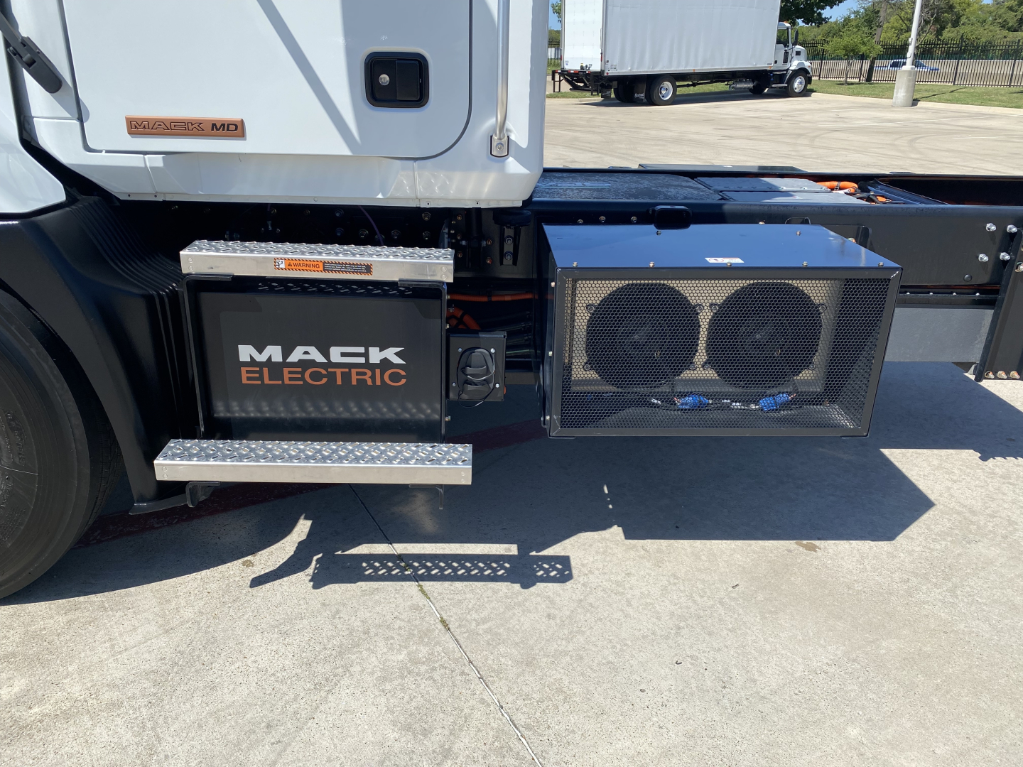 Truck cab and chassis with Mack Electric battery pack mounted, side steps, and radiator/grille area visible on right.
