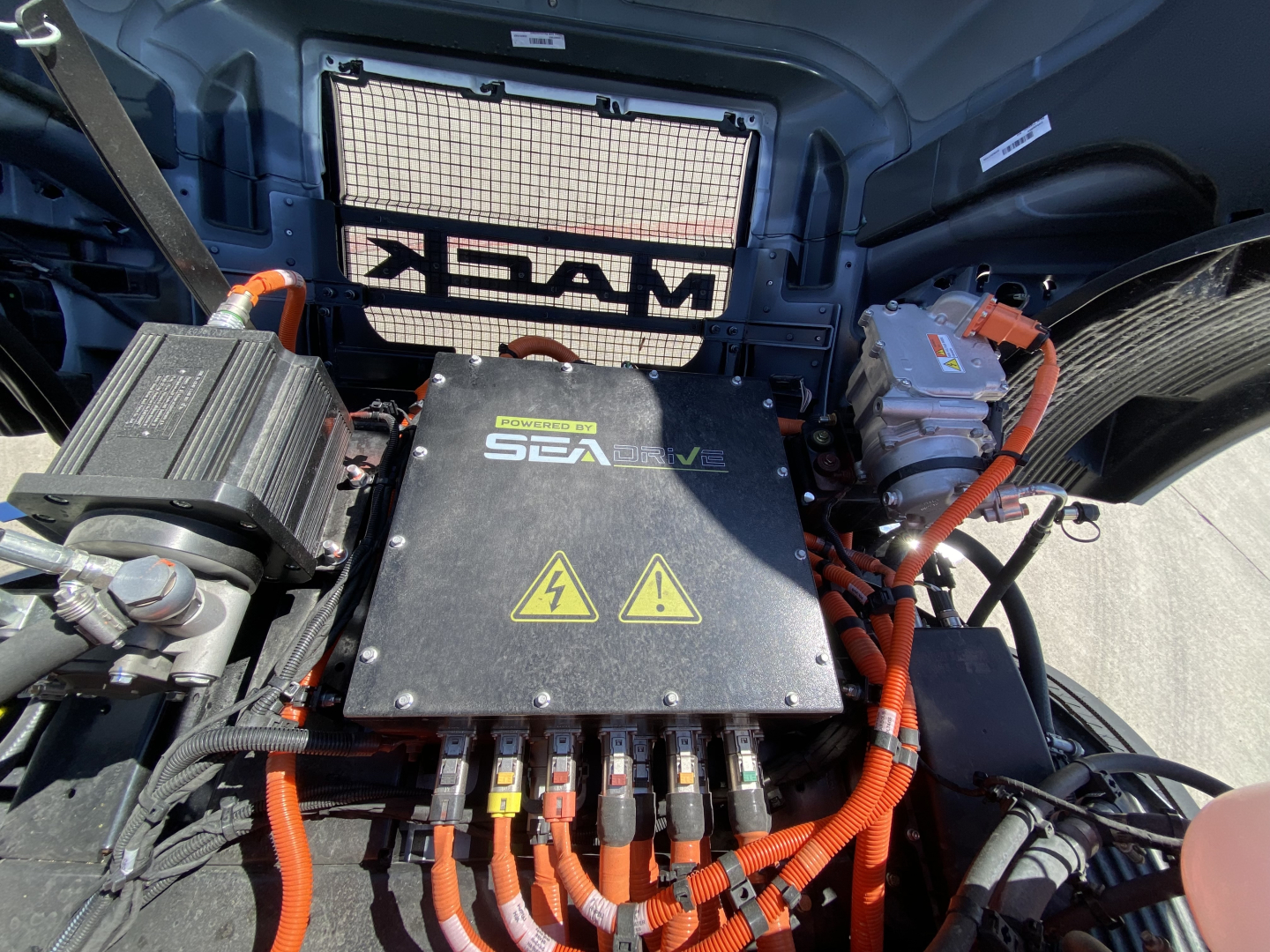 Mack MD6 E cab and chassis engine bay showing SeaDrive hybrid/electric control module with warning symbols, orange high-voltage cables, and battery/aux components.