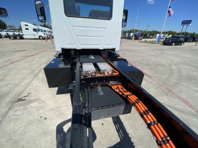 View of a Mack MD6 E cab and chassis with the drive line, battery packs, and orange cables visible during daytime; no legible text on components.