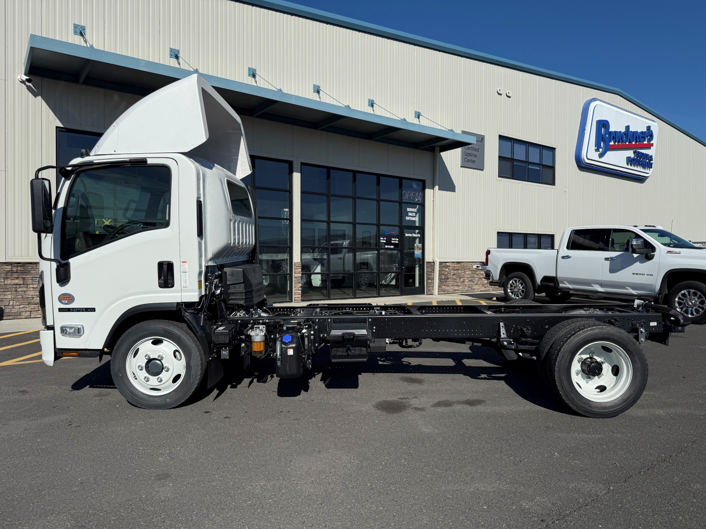Isuzu NPRXD cab and chassis truck in front of dealership building with logo sign in background, white cab, chassis frame visible, side view.
