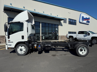 Isuzu NPRXD cab and chassis truck in front of dealership building with logo sign in background, white cab, chassis frame visible, side view.