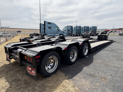 Lowboy trailer with triple-axle rear, Long King brand mudflap visible, 9'9' platform, multiple axles, reflective lights, cab of semi-truck in background.