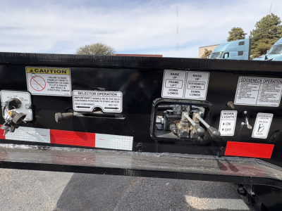 Trailer rear bumper with multiple warning labels and control levers, Gooseneck operation panel, 'CAUTION' and instruction stickers, black painted steel surface.