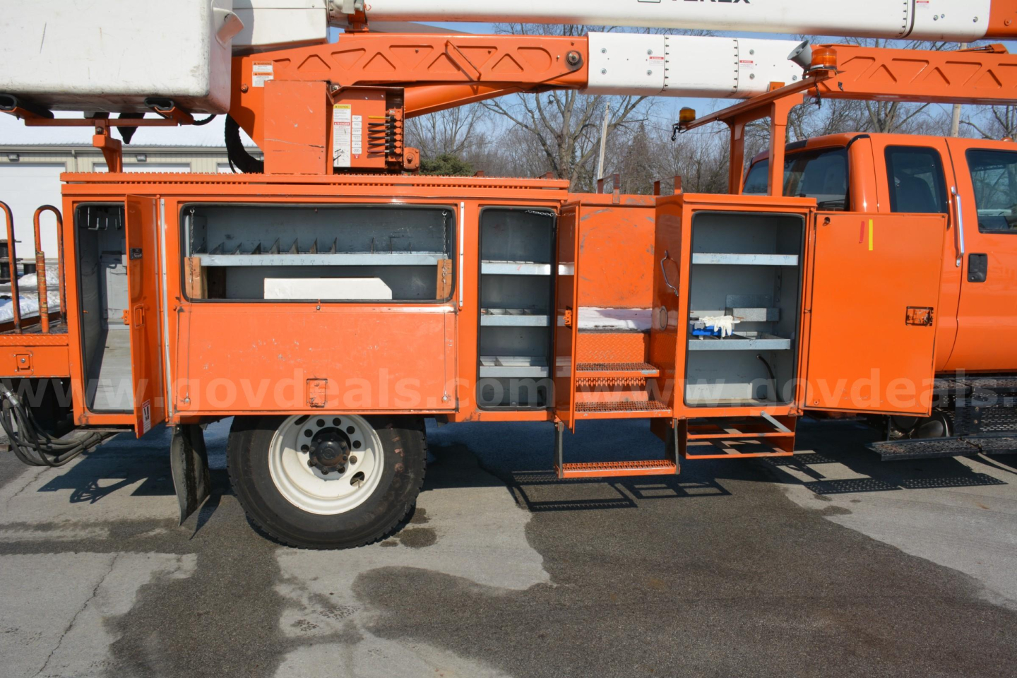 Side view of an orange Ford F-750 bucket truck with open compartments and shelving, showing equipment storage on the passenger side