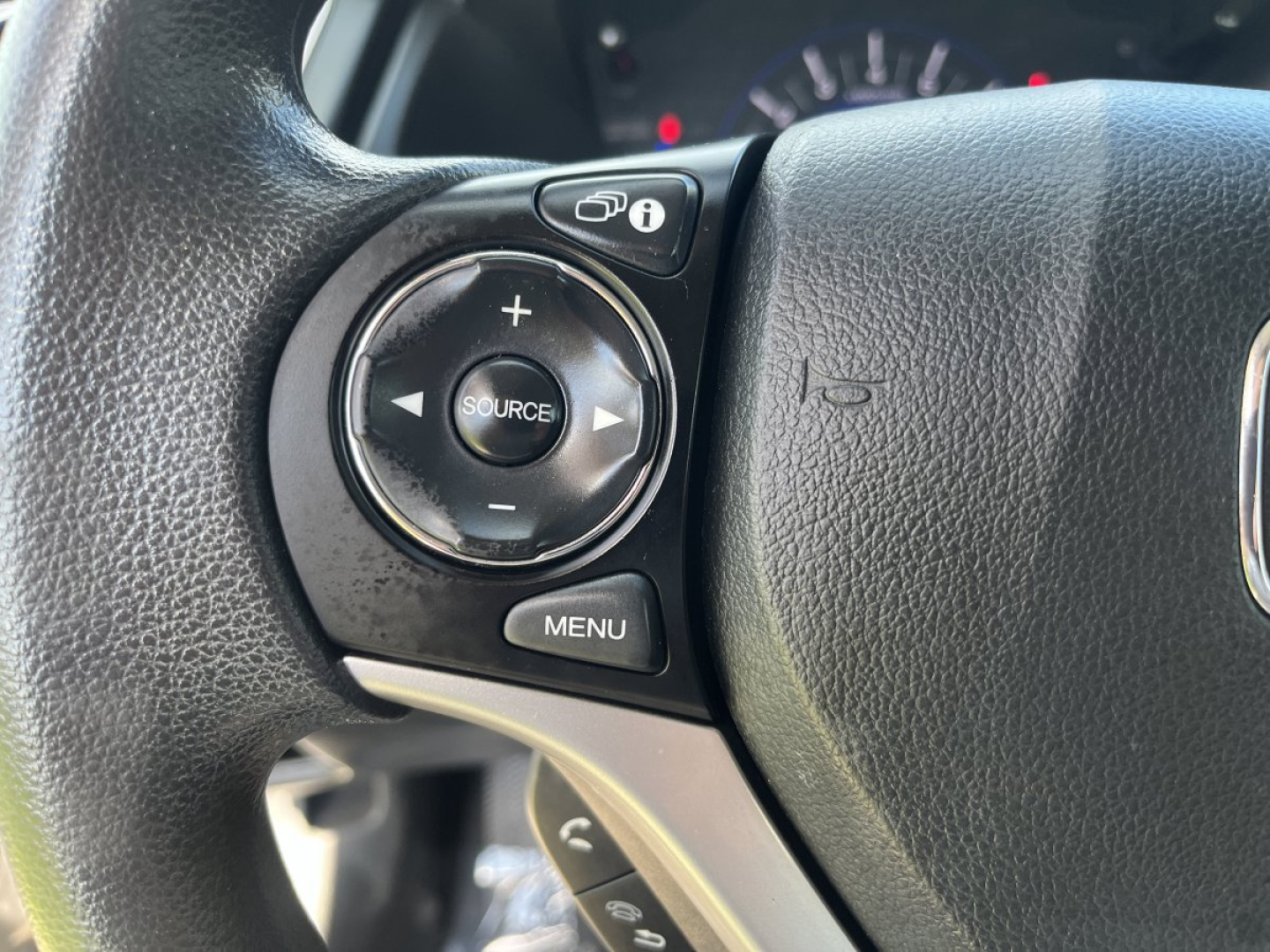 Steering wheel with audio control cluster showing circular pad with + and - buttons and center SOURCE button, plus MENU button and steering wheel logo area.