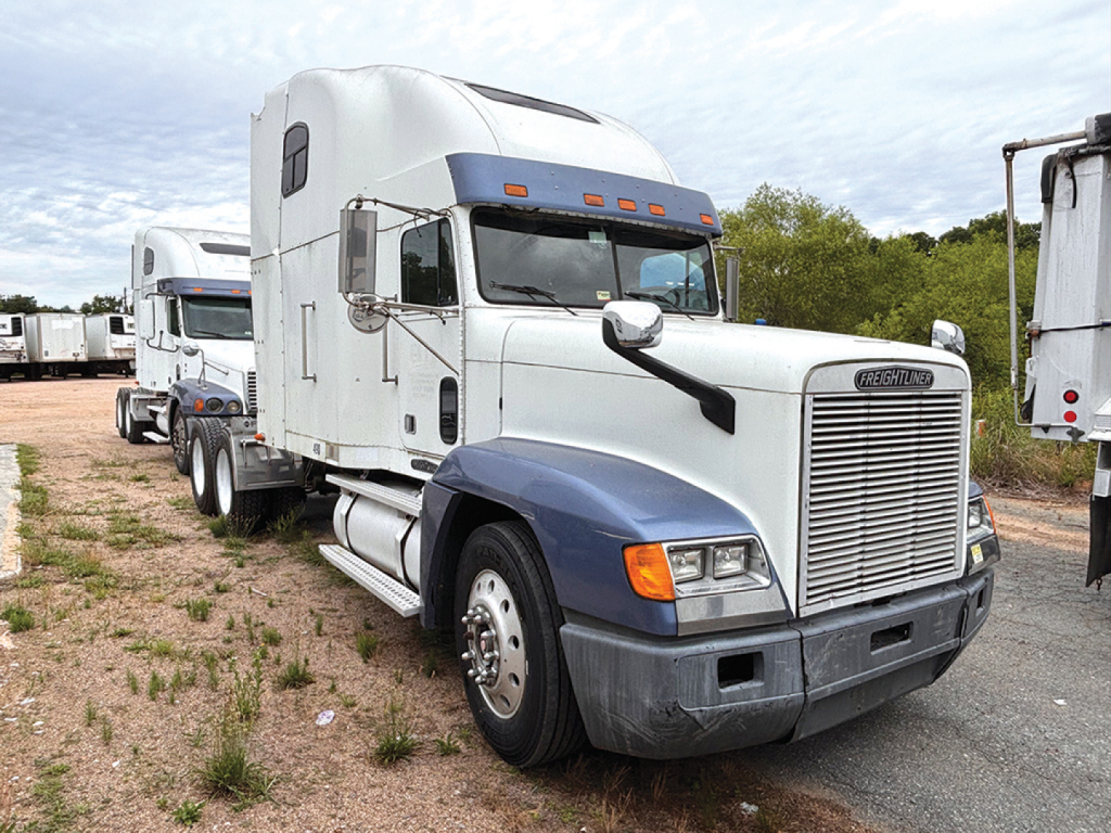 White Freightliner FLD120 truck in a yard, front-right view showing Sleeper, chrome grille, blue lower bumper, large side mirrors, and attached trailer behind.