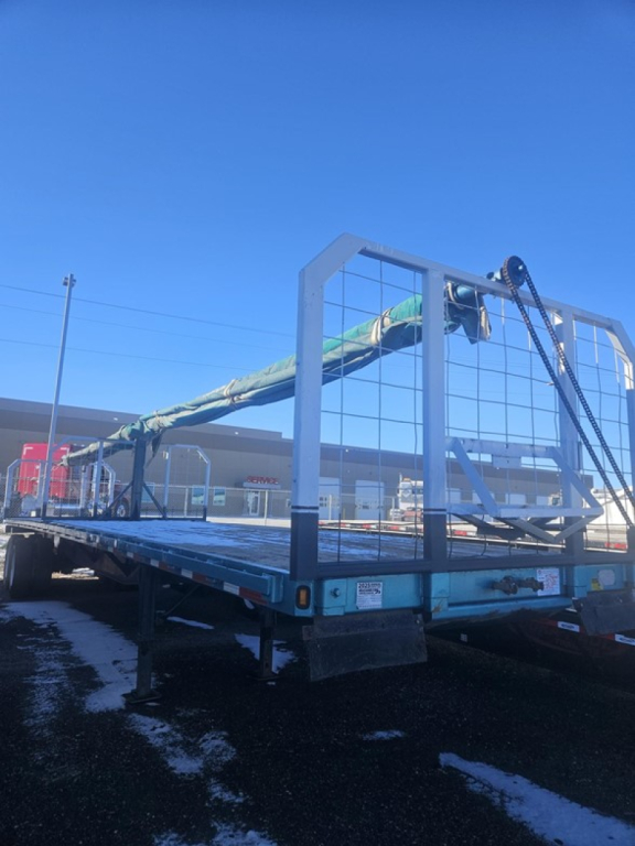 Trailer bed showing blue flatbed with metal mesh gooseneck safety rack and folded side rails, rear view with tires and support legs visible.