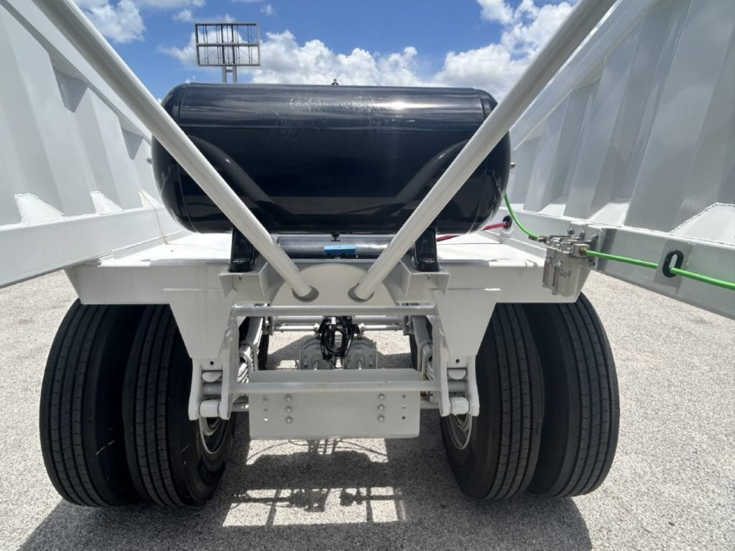 Dump trailer rear view showing dual rear axles, white frame, black cylindrical tank, and safety latch with green strap.