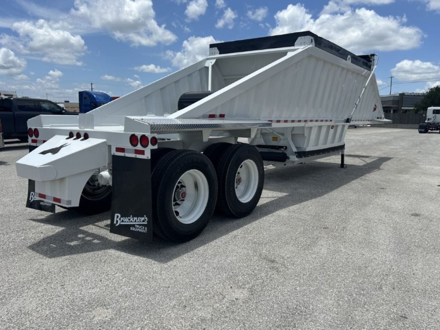 Trailer in service position showing a white dump trailer with dual rear axles and hydraulic system, open-top bed, and support legs visible.