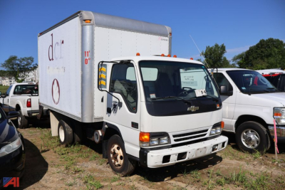 Box truck cab over with white cab and box, Chevrolet badge on grille, side mirrors, and a small cargo box; visible cars in lot.