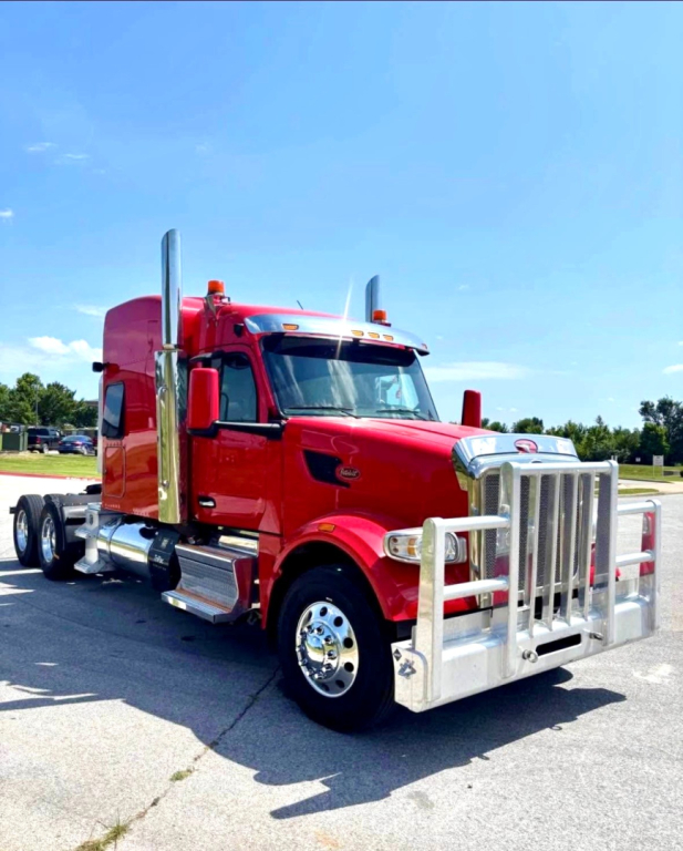 Peterbilt 567 conventional sleeper truck in red with a heavy-duty front bumper and chrome exhaust stacks, side view, clear sky.