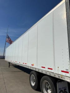 Trailer side view showing white Wabash trailer with dual rear axles, reflective tape, and white hardware; American flag in background.
