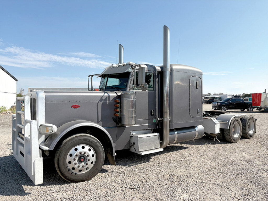 Peterbilt 389 extended hood semi-truck, gray exterior, dual stacked exhausts, large front bumper guard, long chassis with triple rear axles, gravel lot.
