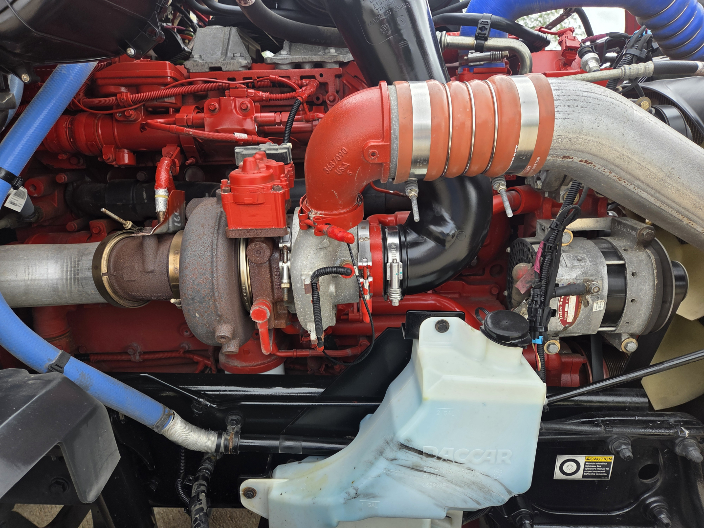 Kenworth T680 engine bay showing red engine components, large orange turbo intake hose, grey intercooler hose, coolant reservoir in light blue, and turbocharger with metal piping.