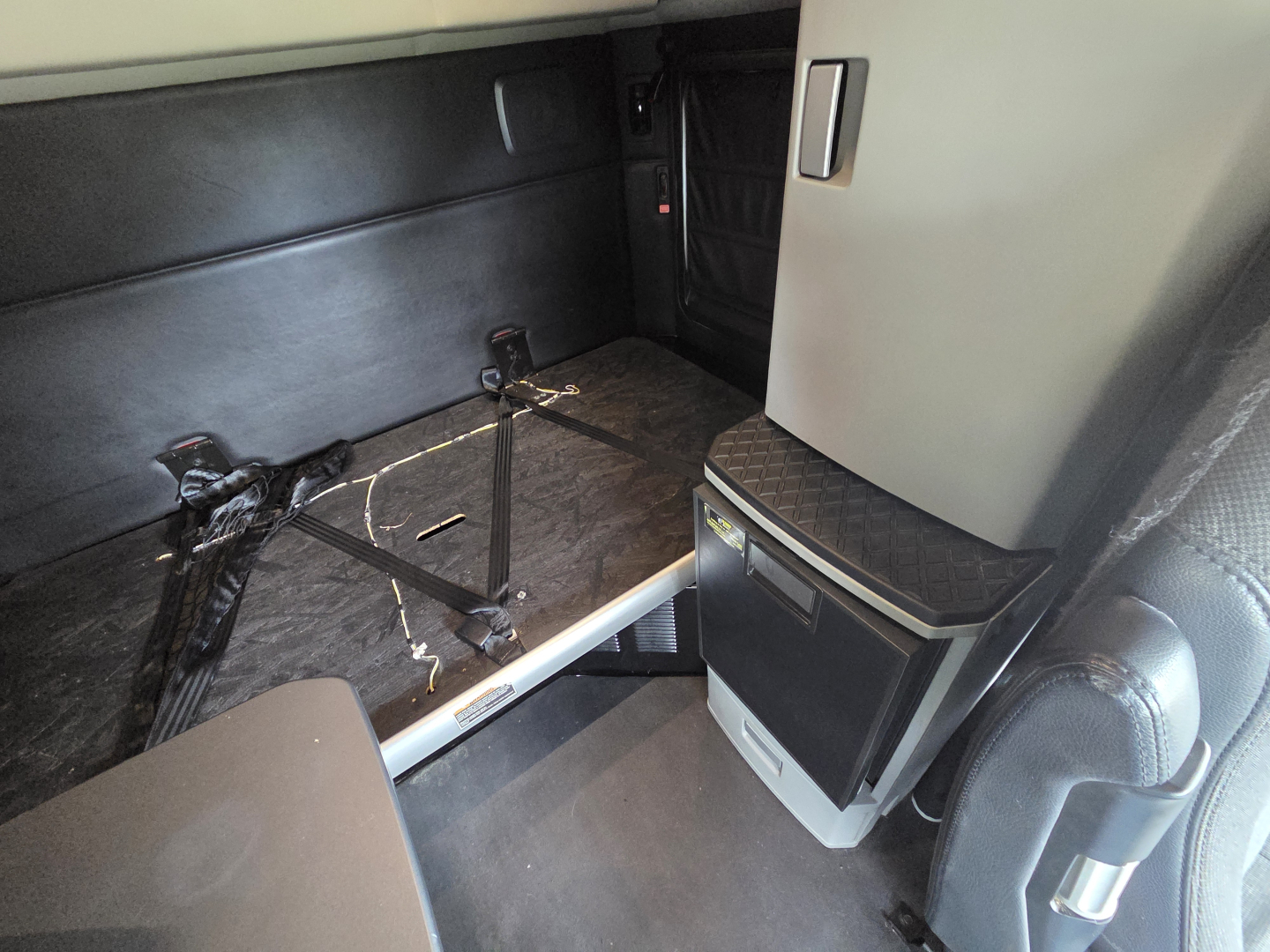Kenworth T680 sleeper cab interior showing seating area and floor with wall panels and a small cabinet; visible cables and seat mounting hardware.