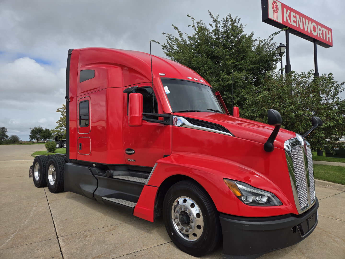 Kenworth T680 semi truck in red, side view showing cab and sleeper with Kenworth sign in background.