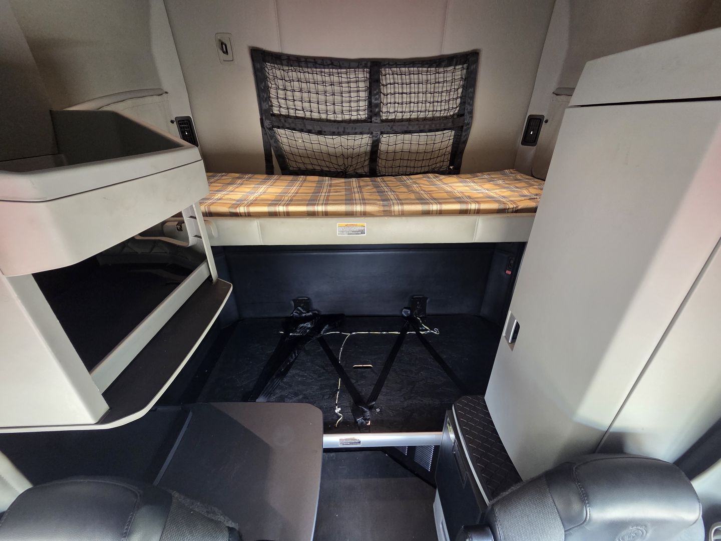 Cab interior of a 2023 Kenworth T680 sleeper cab showing bunk area with plaid cover, storage net, and seating in view.
