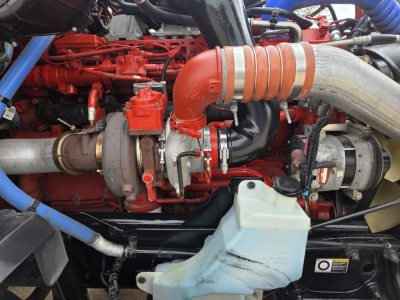 Kenworth T680 engine bay showing red engine components, turbocompressor, large light-orange intake pipe, silver/gray exhaust piping, coolant reservoir, and various hoses.