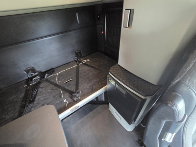 Truck interior rear sleeper area with mounting brackets and cables visible on floor, side cabinet and door visible in cabin interior.