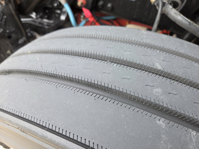 Tire tread close-up on a Kenworth T680 front axle showing rubber tire with visible wear patterns and surrounding engine bay components.