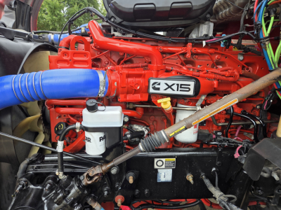 Kenworth T680 engine bay with bright red X15 engine and blue intake hose, visible fluid reservoirs, wiring, and components in a sleeper truck.