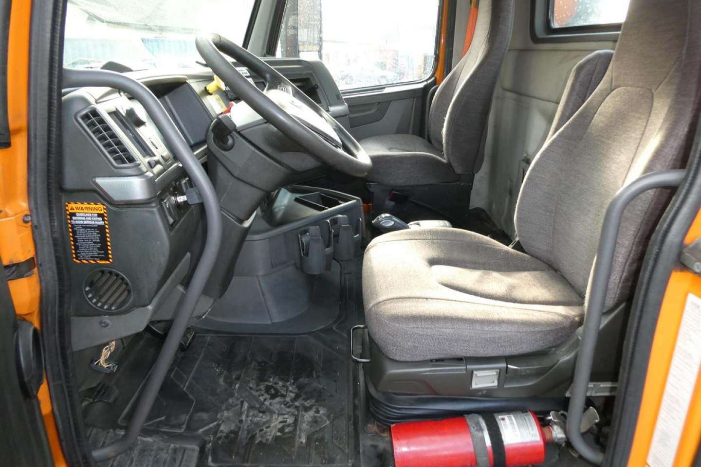 Truck cab interior showing steering wheel, driver seat, and passenger seat with gray fabric. Dashboard and pedals visible; orange exterior edge; floor mat and a red fire extinguisher at floor near seat.