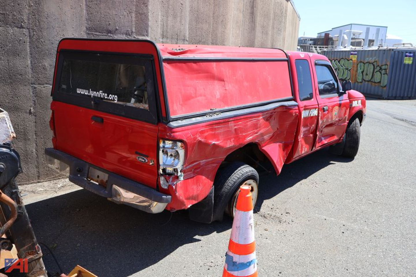 Ford Ranger pickup with extended cab and cargo cap, red paint, dented side, rear view showing tailgate and bumper, in a lot with an orange traffic cone.
