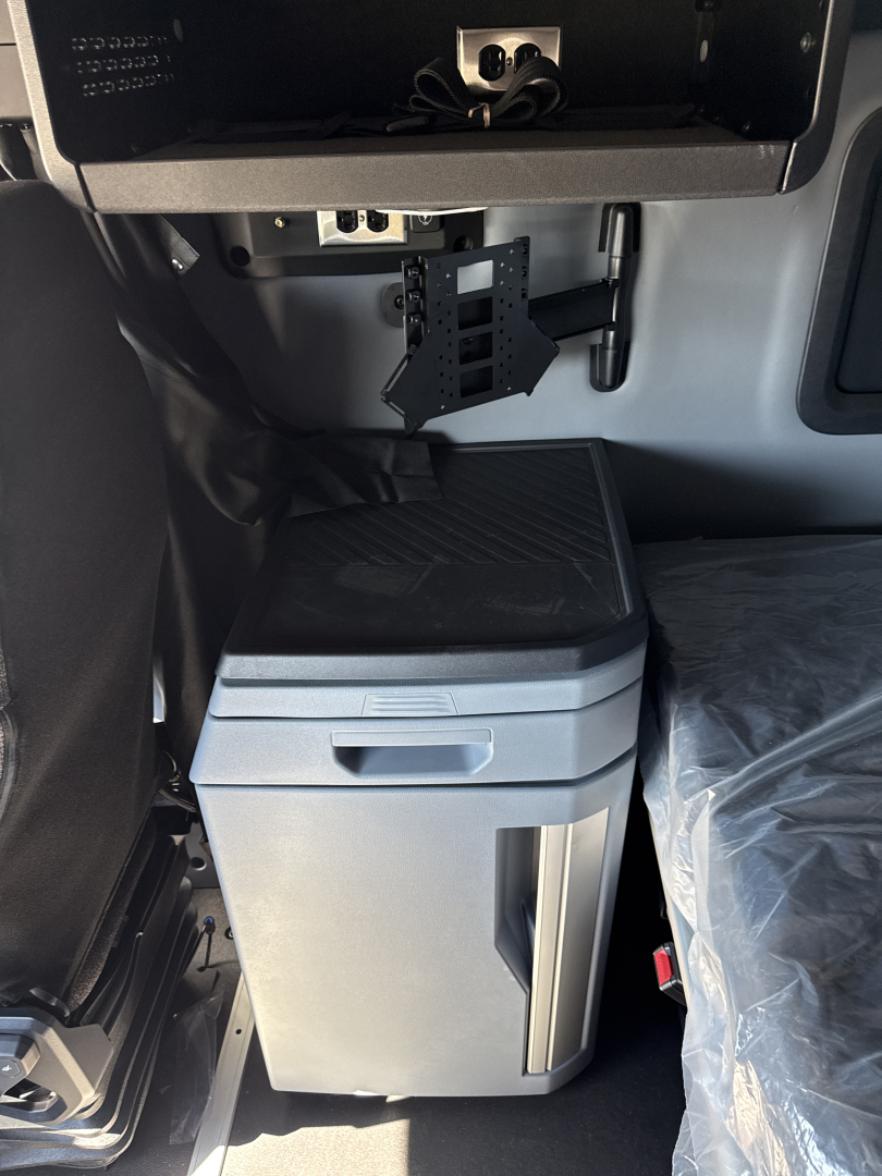Cab interior of a 2026 Mack PR64T76H showing a storage cabinet/refrigerator under the dash and a hanging electronics mount with cables.