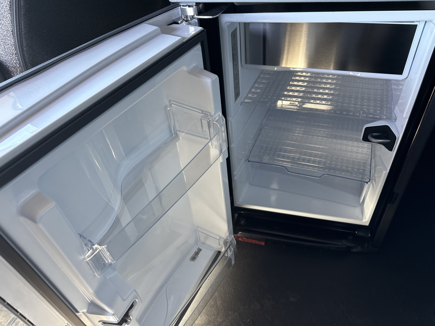Conventional sleeper truck dash area? image shows an open small refrigerator/freezer unit with empty shelves located in a storage compartment; door on left with shelves visible; right side shows another small fridge/freezer compartment.