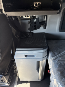 Cab interior of a 2026 Mack PR64T76H showing a storage cabinet/refrigerator under the dash and a hanging electronics mount with cables.