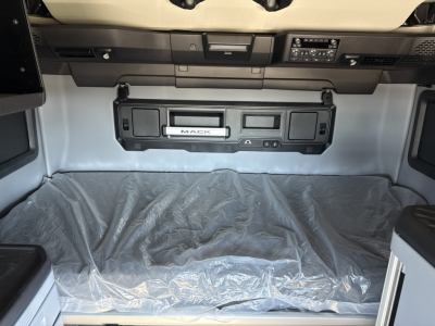 Truck cab interior showing Mack logo panel and dashboard storage above the sleeper area; plastic cover on the floor ahead of storage compartment.