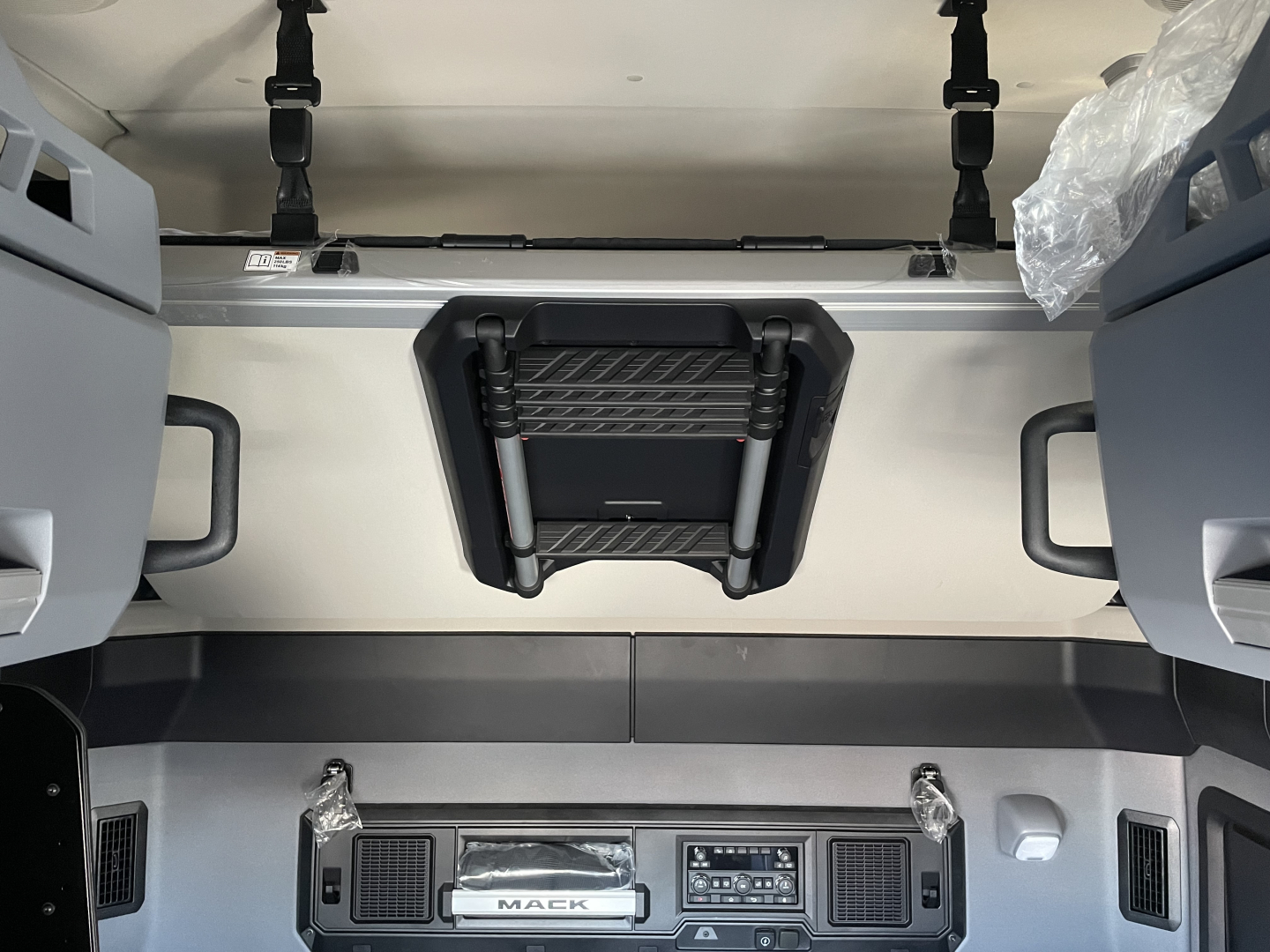 Interior view of a new Mack PR64T76H truck cab showing overhead sleeper access, dash controls, and seating mounts.