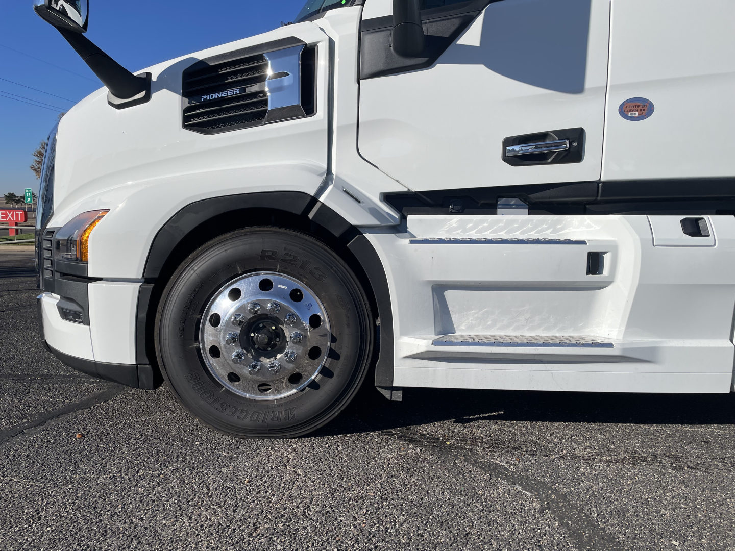 Truck wheel and front fender of a white Mack PR64T76H sleeper cab showing chrome hubcap and side step tread plate.