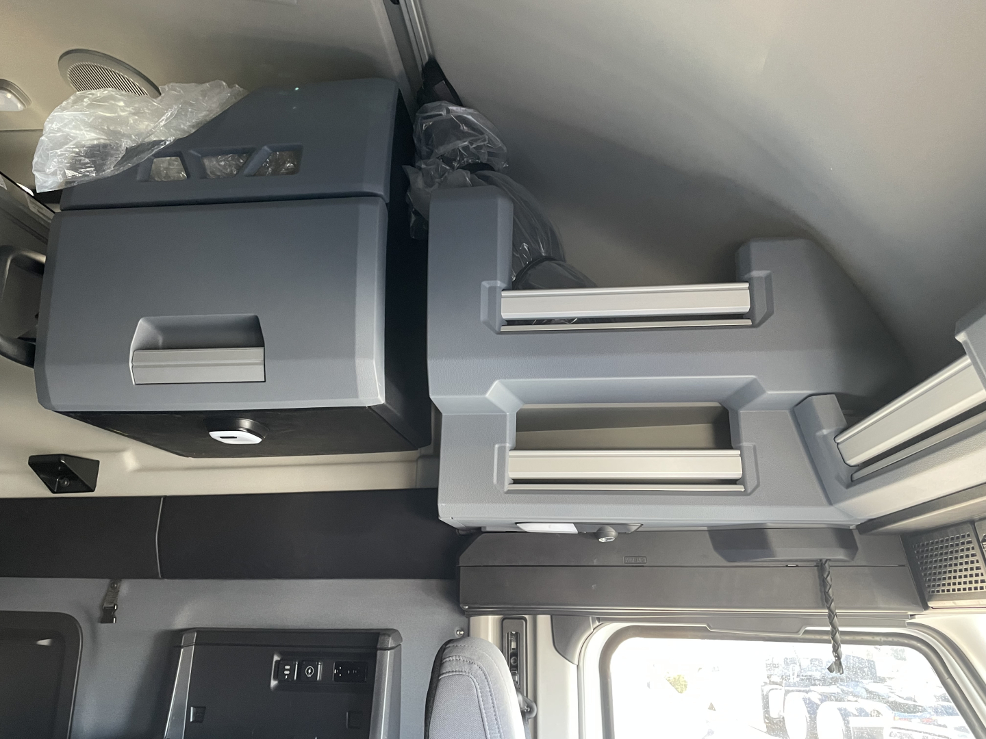 View inside a Mack PR64T76H sleeper cab showing overhead storage with drawers and compartments, a gray console, and a window; no legible text visible.