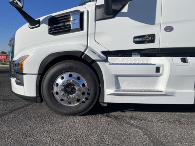 Truck wheel and front fender of a white Mack PR64T76H sleeper cab showing chrome hubcap and side step tread plate.