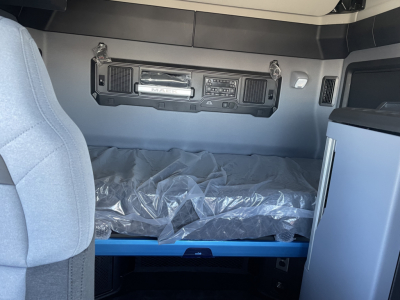 Mack PR64T76H truck cab interior showing overhead control panel, storage bunk with plastic-wrapped mattress, and blue bed frame.