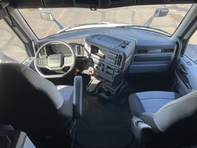 Dashboard and steering wheel of a Mack truck interior showing the driver seat, steering wheel with Mack logo, central control console with various buttons, gear shifter, and passenger seat.