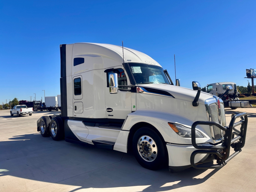 Truck image: white 2023 Kenworth T680 conventional sleeper, front bull bar, large grille, side mirrors, dual rear axles, sleeper cab, clear sky.