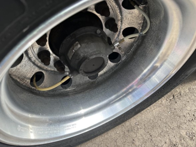 Trailer wheel and hub showing steel rim, black ABS/parking sensor component, and hoses; close-up of hub area with tire tread visible.