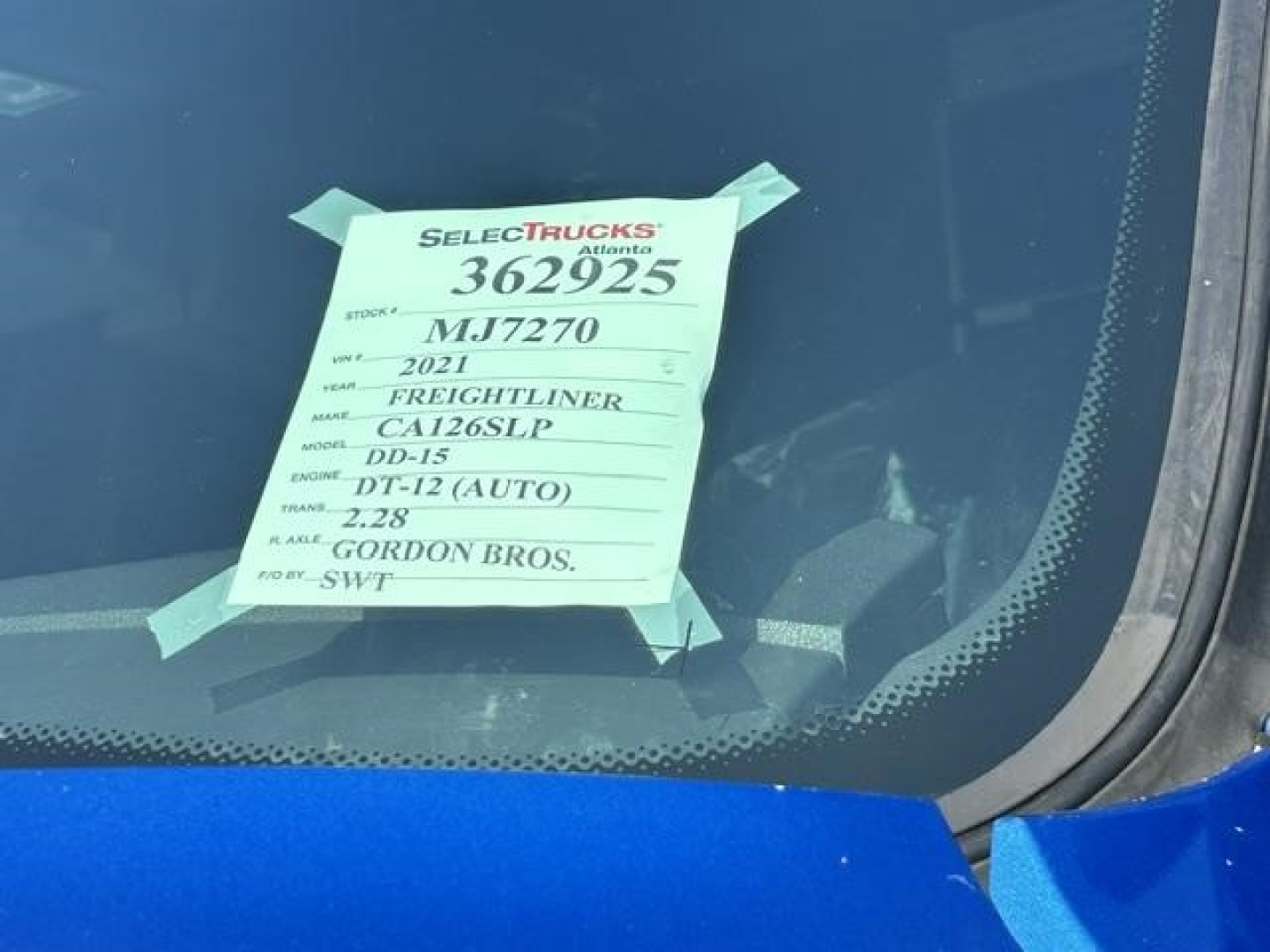 Truck window with a white listing tag taped to the glass showing serial numbers and model codes for a 2021 Freightliner Cascadia 126. Text includes 362925, MJ7270, 2021, FREIGHTLINER, CA126SLP, DD-15, DT-12 (AUTO), 2.28, GORDON BROS.