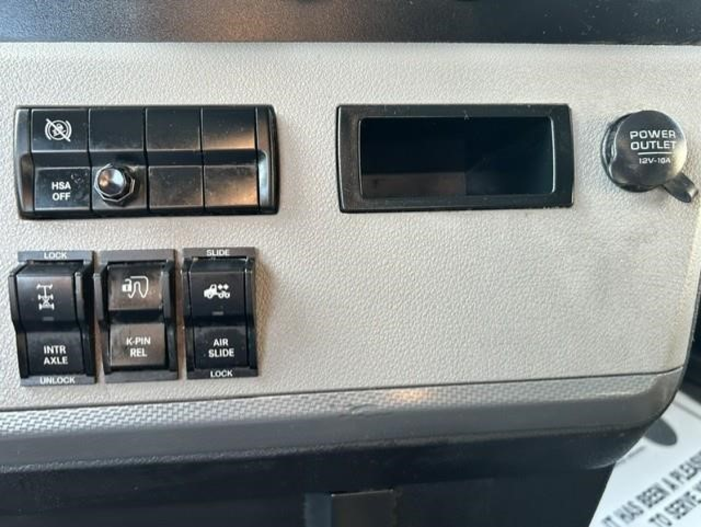 Cabin panel of Freightliner Cascadia showing switches and ports, including HSA OFF, INTR AXLE, K-PIN REL, AIR SLIDE, power outlet, blank slot, and miscellaneous control buttons.