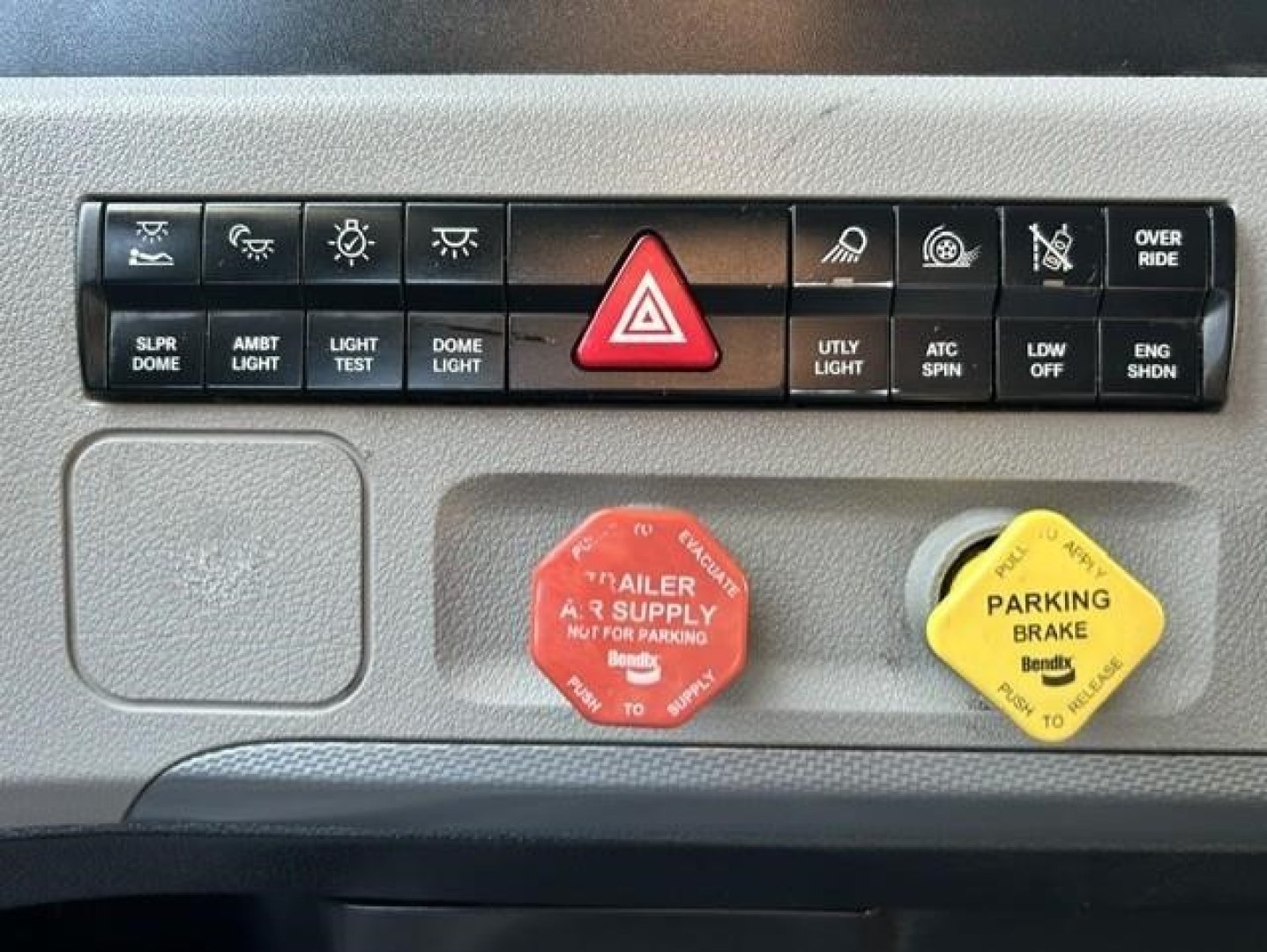 Truck dash panel showing red trailer supply honk cap and yellow parking brake cap with hazard button in center and various control buttons above.
