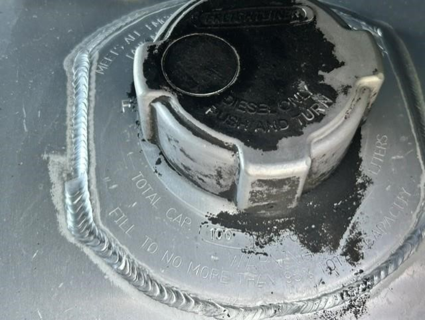 Fuel tank cap and surrounding fuel filler area of a Freightliner Cascadia. Visible are the circular cap with locking ring, fuel filler neck, and embossed text around the cap including ‘FILL TO NO MORE THAN’ and other maintenance marking