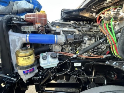 Engine bay of a Freightliner truck showing blue intercooler hose, yellow coolant reservoir cap, translucent coolant overflow bottle, and various wiring harnesses with colored cables.