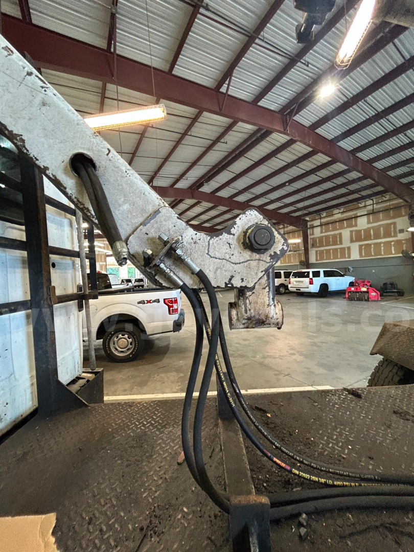 Boom truck boom arm with hydraulic hoses and pivot point in foreground; warehouse interior in background with parked vehicles.
