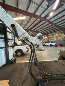 Boom truck boom arm with hydraulic hoses and pivot point in foreground; warehouse interior in background with parked vehicles.