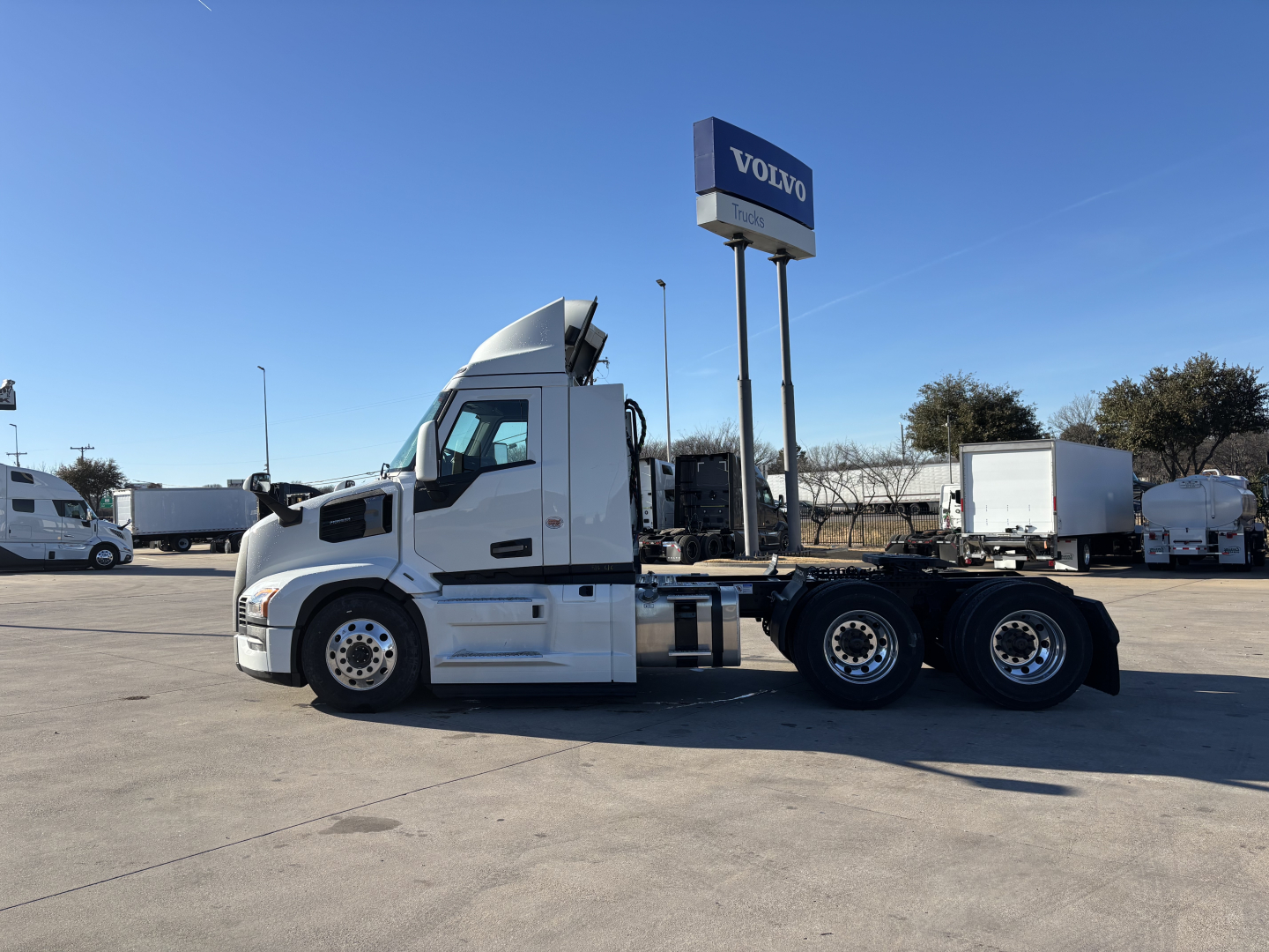 White 2027 Mack PR64T truck tractor side view in a dealership lot with a Volvo sign in background.