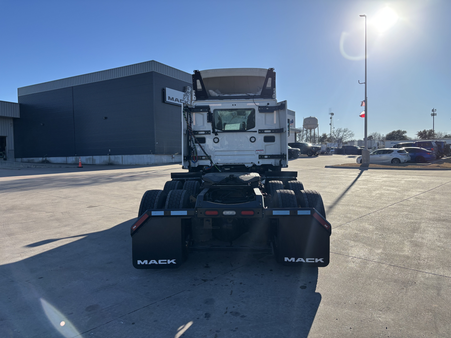 Front view of a 2027 Mack PR64T truck chassis in a dealership lot, showing dual rear wheels, mud flaps with 'MACK' branding, and cab at distance.