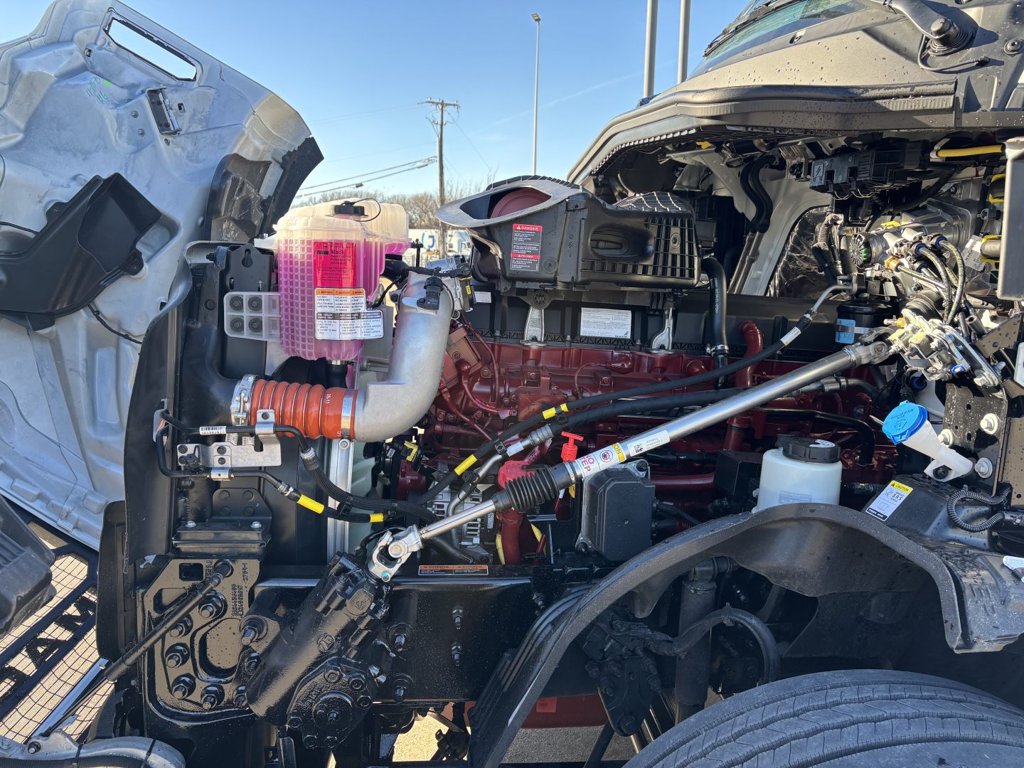 Engine bay of a 2027 Mack PR64T truck showing red engine block, intake manifold, hoses, and various components.