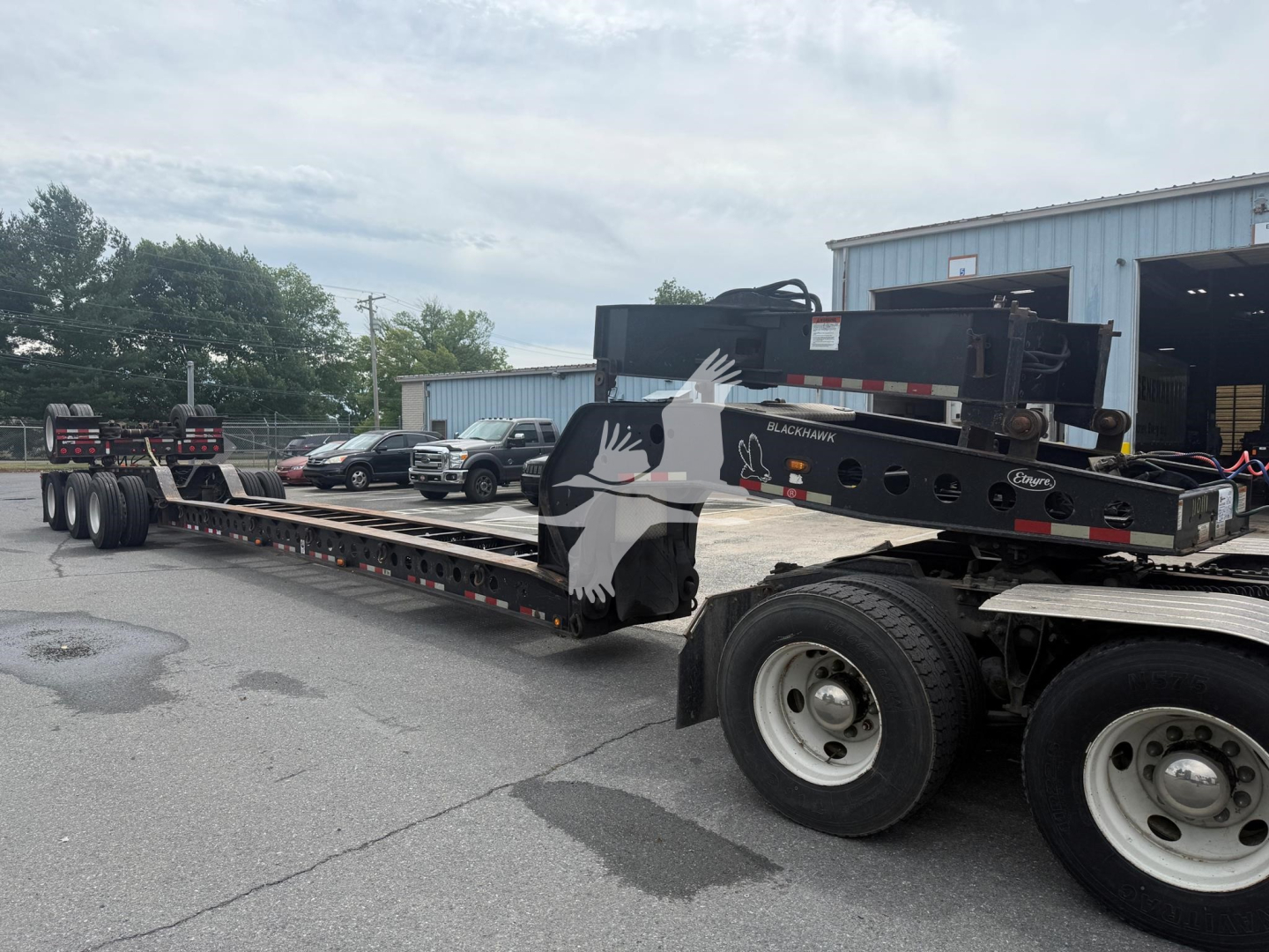 Etnyre beam lowboy trailer showing long flat deck and dual rear axles with black crane attachment and bird graphic on side.